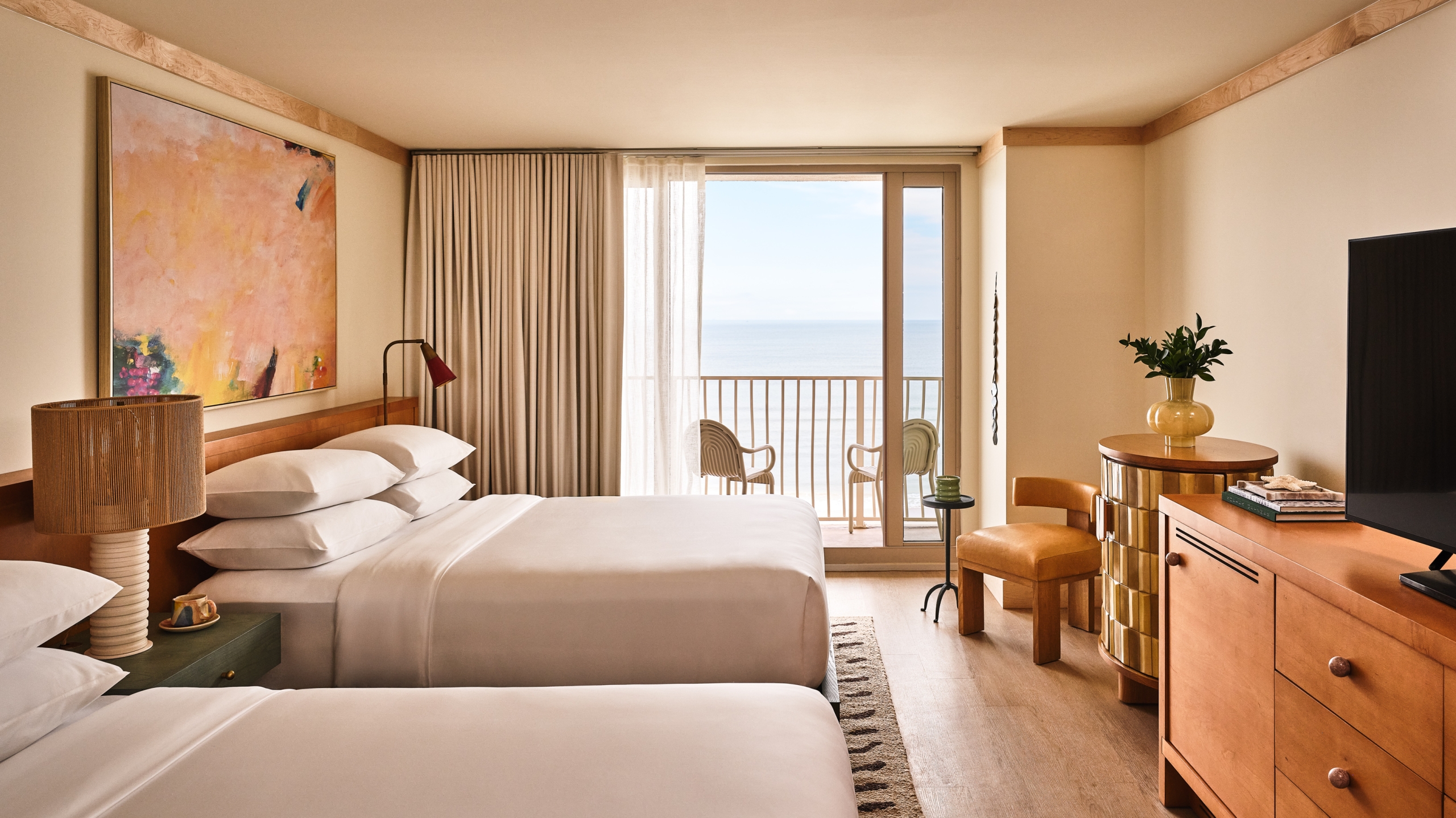 A bright hotel room with two queen beds, abstract art, and a balcony overlooking the blue ocean.