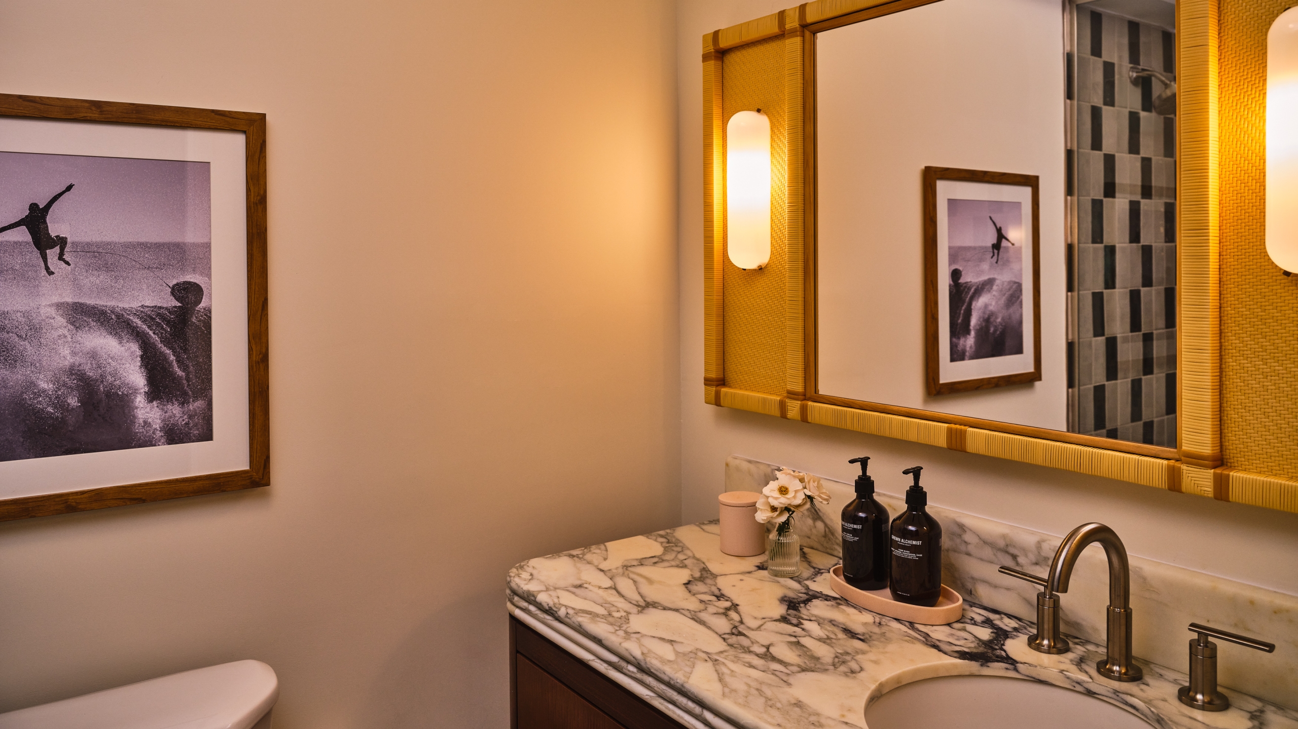 Luxury hotel bathroom with marble vanity, rattan mirror, brass fittings, and black-and-white surf photography on warm beige walls.