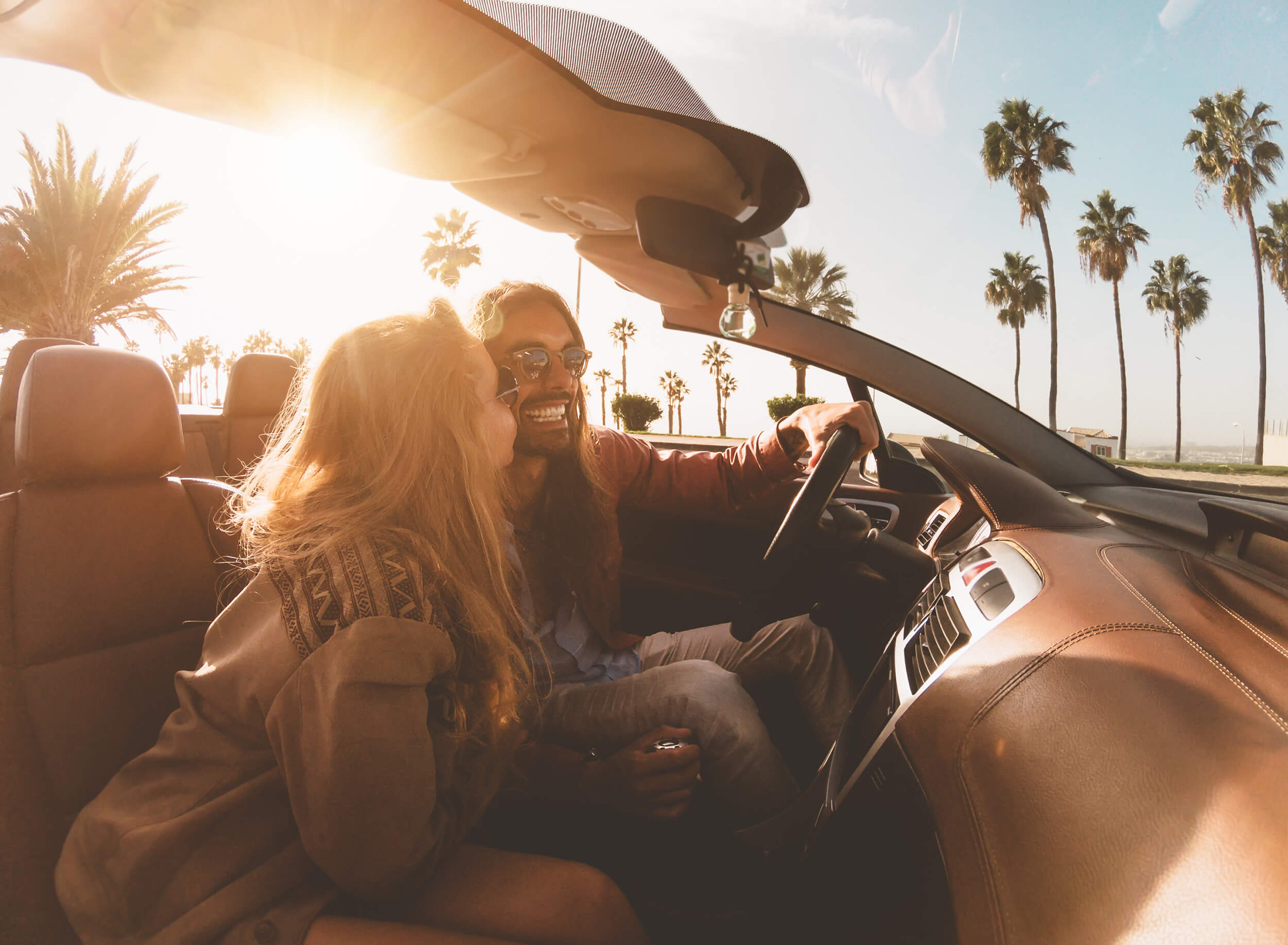 A smiling couple in a convertible car driving past palm trees during a golden sunset.