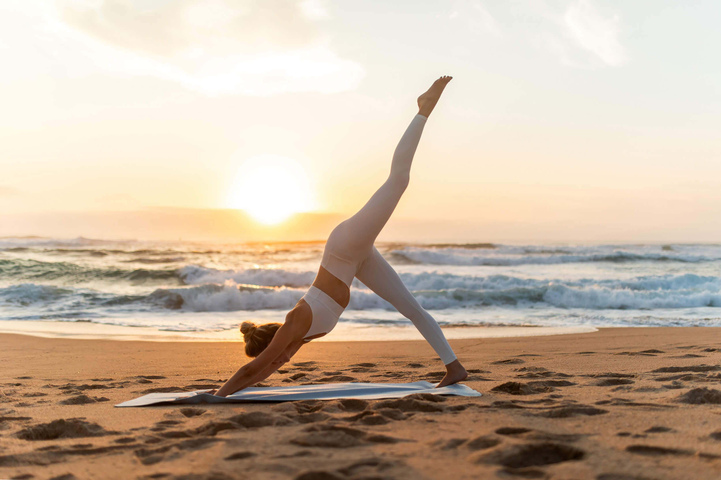 A person in white activewear performing a three-legged downward dog yoga pose on a beach at sunrise.