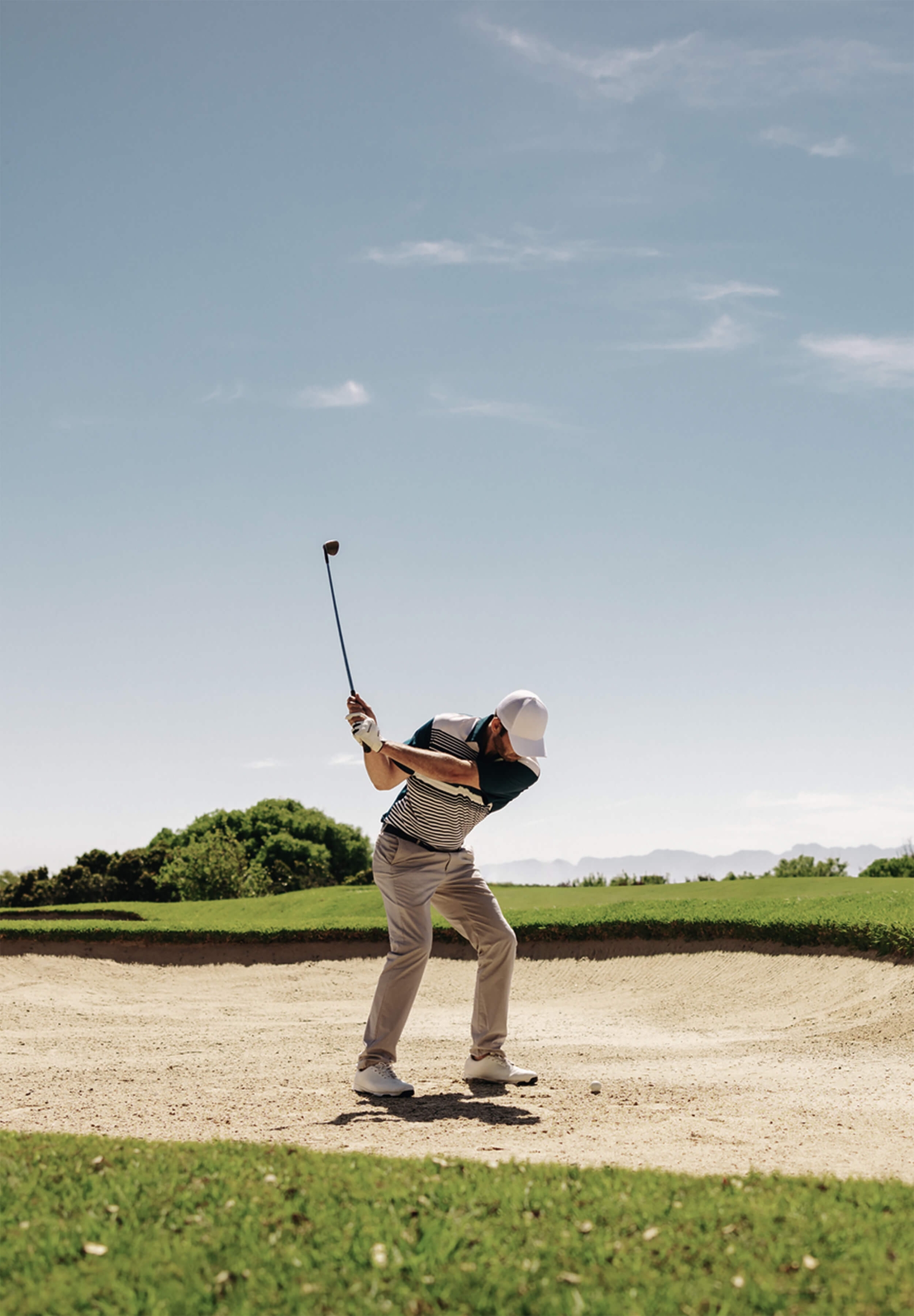 A male golfer in a striped shirt and white cap mid-swing in a sand trap under a clear sky.