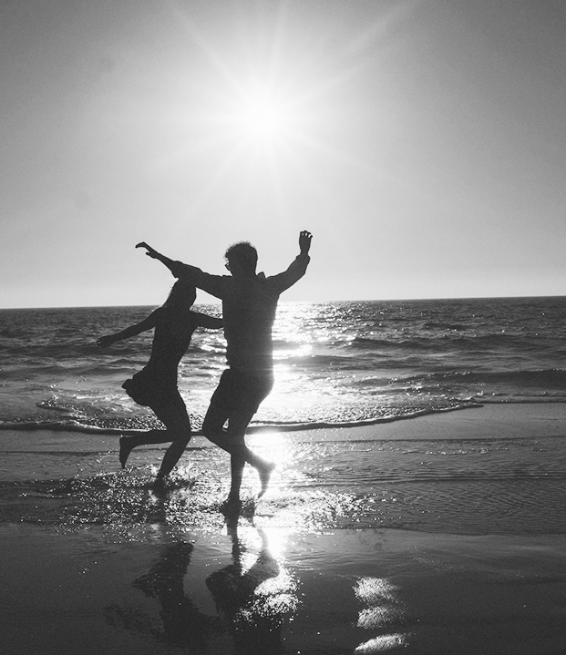 Black and white silhouette of a couple joyfully dancing in the shallow waves of a beach at sunset.