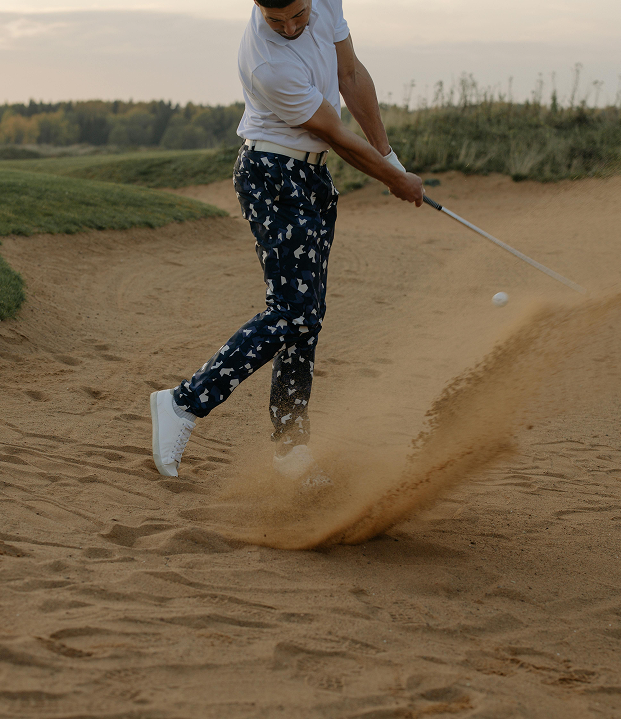 A golfer in navy patterned trousers swings a club in a sand bunker, sending a spray of sand into the air.