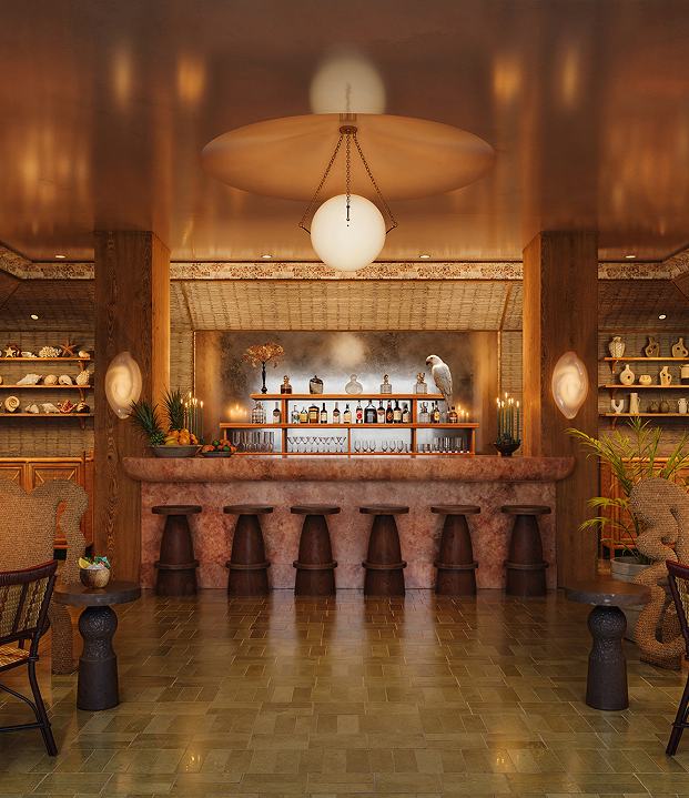 A luxury hotel bar with a pink marble counter, dark wood stools, and shelves filled with curated coastal decor.