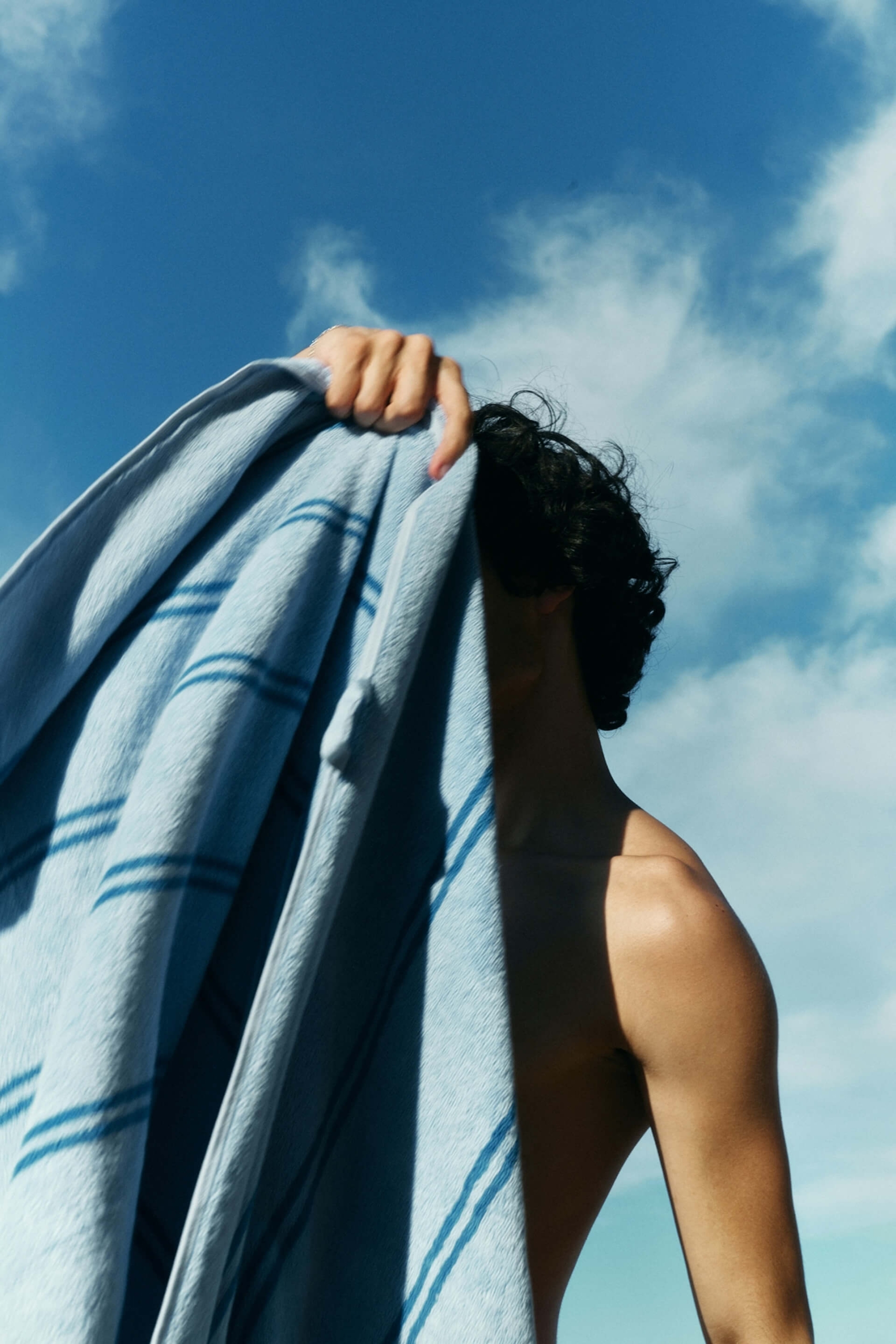 A person holding a blue striped towel up against a bright blue sky with light clouds.