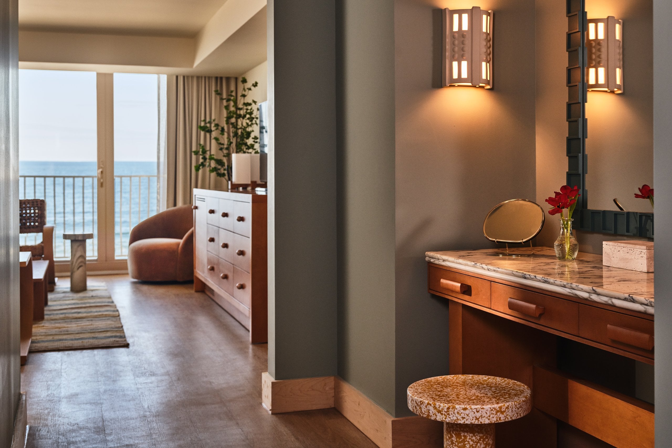 Luxury hotel suite vanity with marble top and brass mirror leading to a sunlit oceanfront living area with balcony.