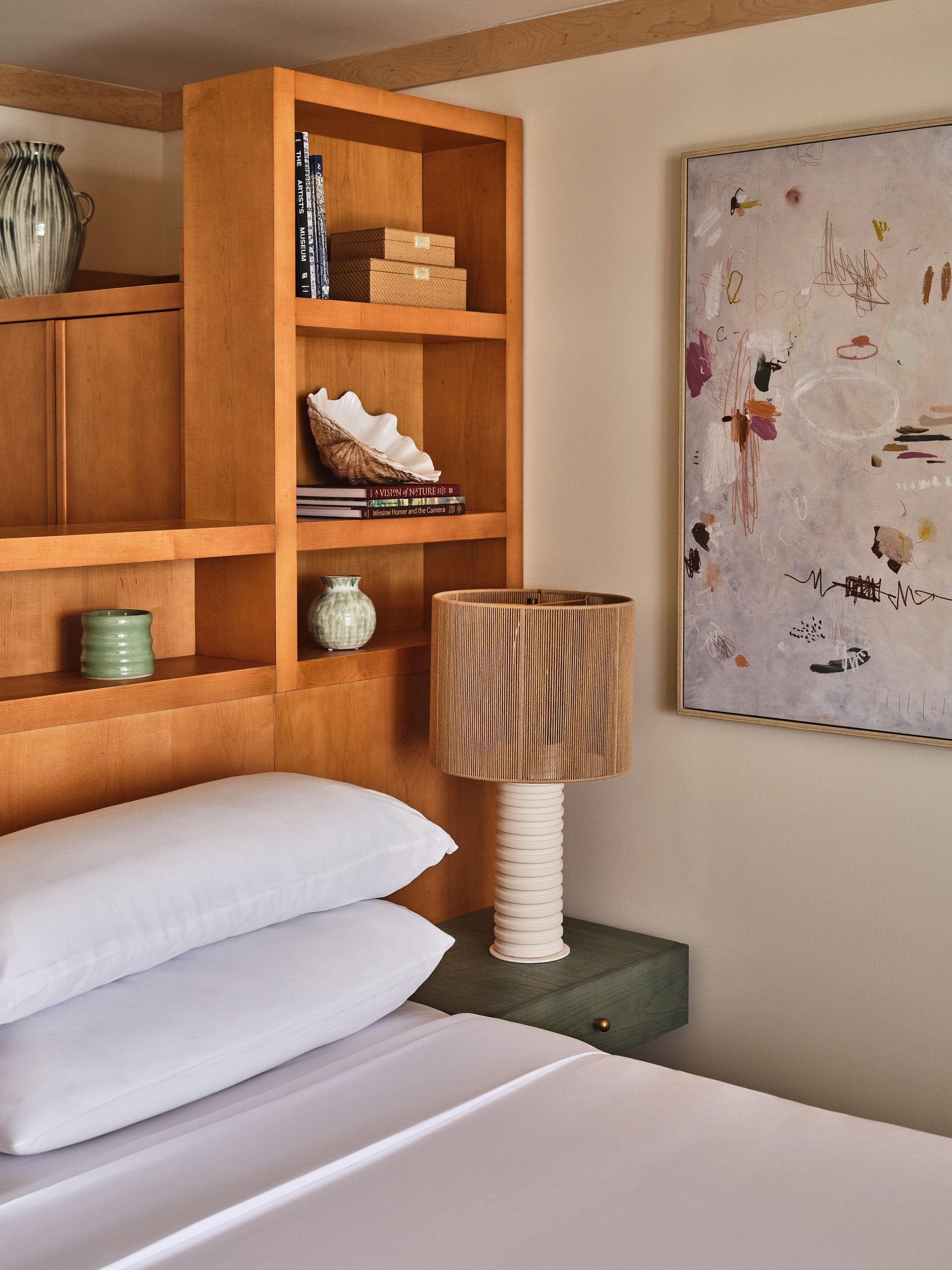 Luxury hotel bedroom with warm wood built-in shelving, ceramic lamp, white linens, and abstract wall art.