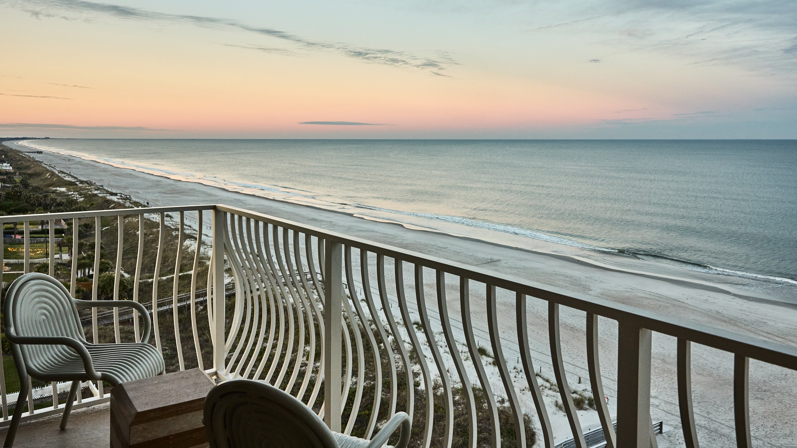 Luxury hotel balcony with curved white railing overlooking a deserted white sand beach and calm ocean at twilight.