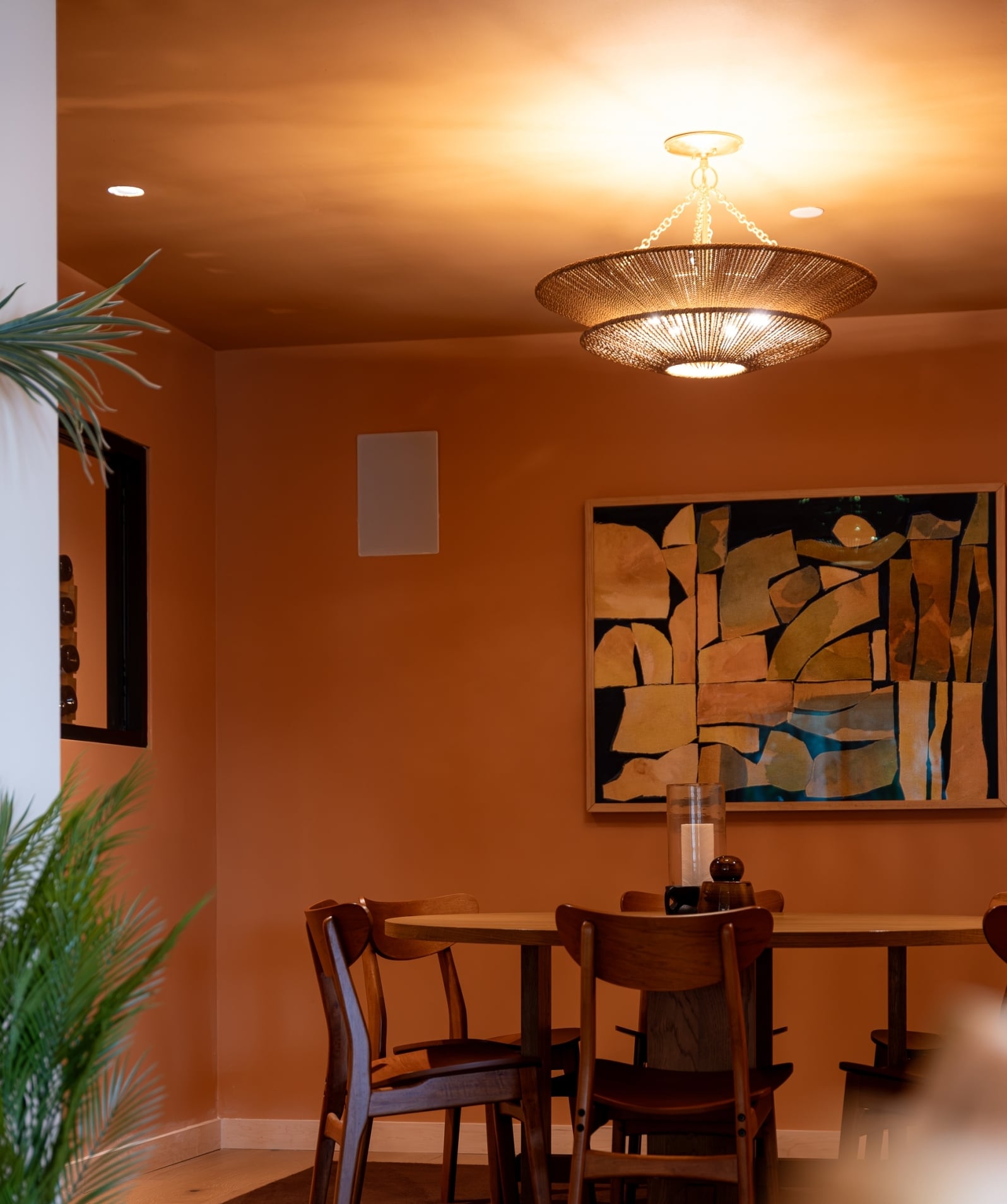 Intimate dining space featuring warm terracotta walls, a wooden dining table, vintage-inspired chairs, abstract artwork, and a woven pendant light
